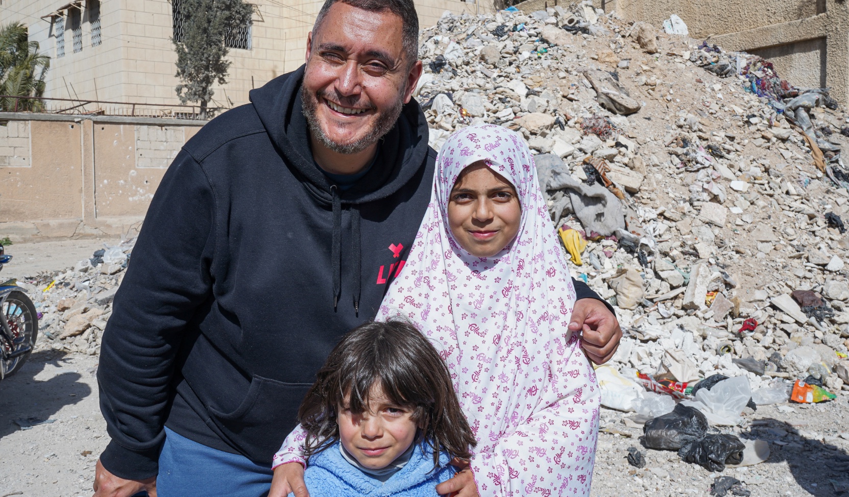 Homme souriant portant un sweat LIFE, entouré de deux enfants, devant un tas de gravats dans une rue.
