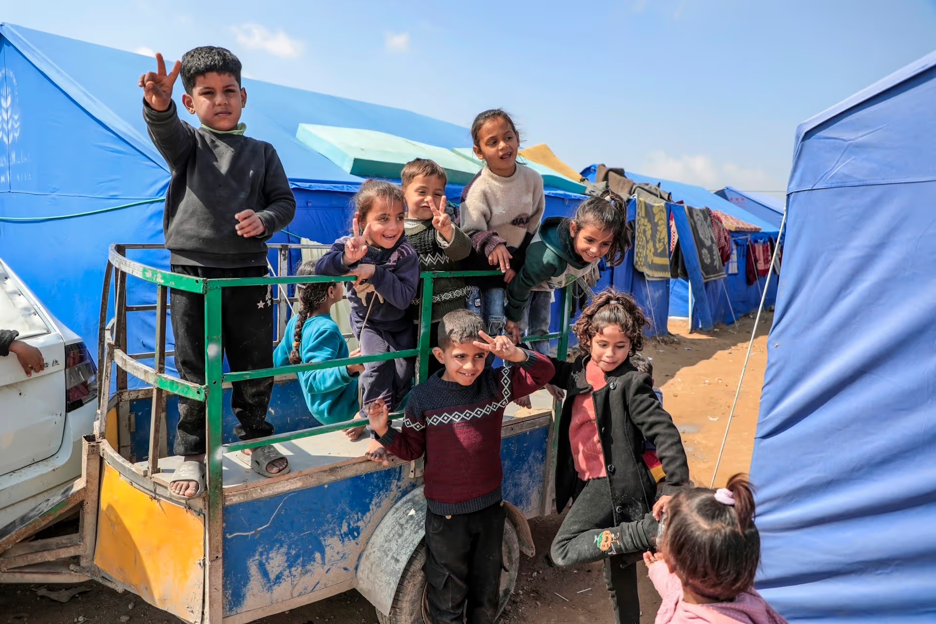 Des enfants sourient et font le signe de la victoire sur une carriole, au milieu des tentes d'un camp de déplacés à Gaza.