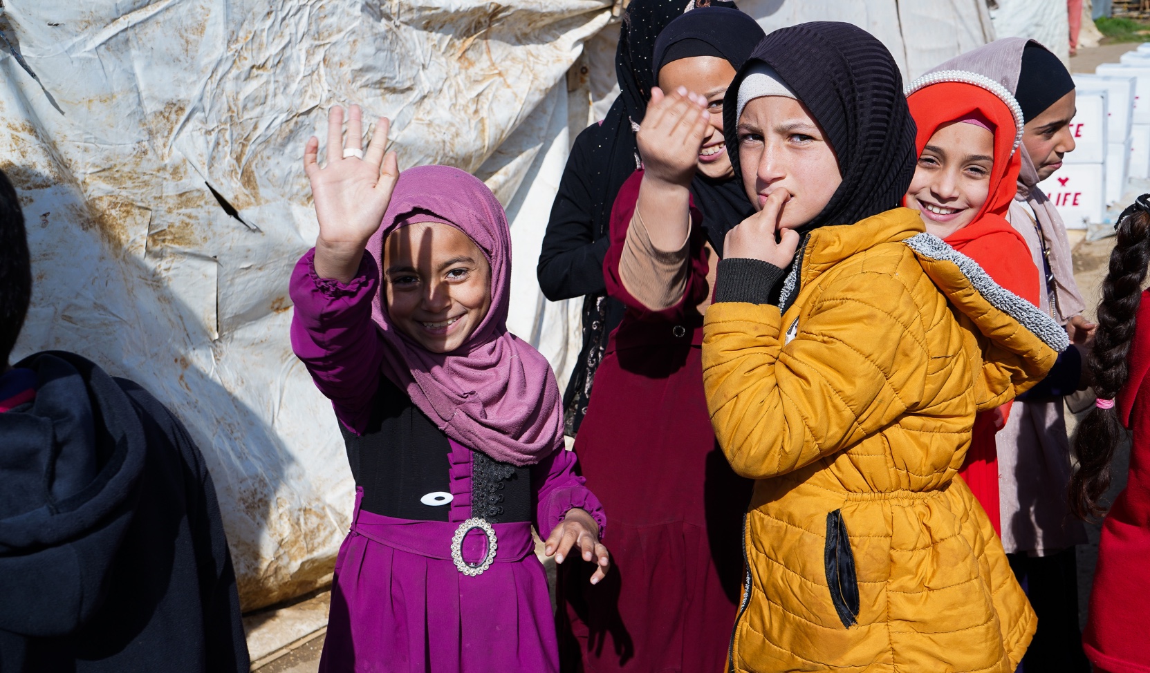 Fillettes souriantes en hijab et manteaux colorés, l'une faisant un signe de la main, devant une tente en bâche usée.